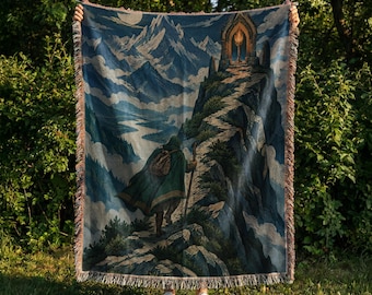 May include: A woven throw blanket featuring a detailed illustration of a hooded figure in a green cloak walking up a rocky mountain path toward a glowing stone archway, set against a backdrop of snow-capped peaks and a deep blue sky.