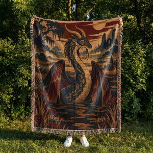 May include: A woven throw blanket featuring a large, central dragon with curved horns and wings spread wide. The scene depicts a swampy landscape with dark blue, brown, and orange tones, including a full moon, trees, and water reflections.