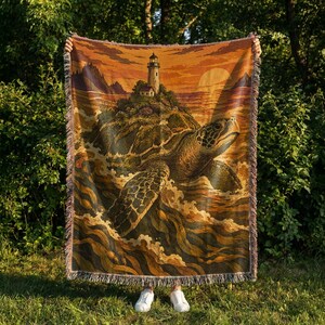 May include: A woven throw blanket featuring a detailed illustration of a large sea turtle swimming through ocean waves toward a rocky island topped with a lighthouse. The scene is rendered in warm shades of orange, brown, and gold with fringed edges.