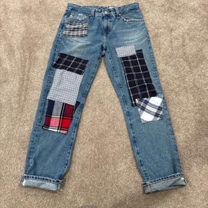 Op de afbeelding: Lichtblauwe spijkerbroek van denim met meerdere rechthoekige stoffen patches in diverse ruitpatronen. De patches in marineblauw, wit, rood en grijstinten zijn op de voorzijde van de broekspijpen genaaid. De jeans heeft een sluiting met knopen.