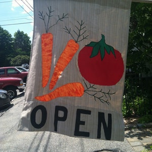 May include: A rectangular fabric banner with a light beige background. The banner features orange carrots and a red tomato with green leaves. The word "OPEN" is displayed in large, black letters. The banner is hanging outside.