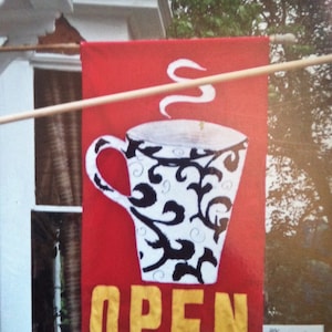 Könnte beinhalten: Eine rechteckige, rote vertikale Flagge mit einer großen weißen Kaffeetasse mit schwarzem Wirbelmuster. Über der Tasse befindet sich eine weiße Dampfgrafik. Das Wort OPEN ist in großen gelben Blockbuchstaben am unteren Rand des Banners gedruckt.