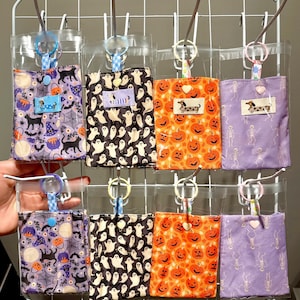 May include: Eight rectangular fabric pouches in clear plastic sleeves, each featuring a key ring and a small rectangular label with a dachshund illustration. Patterns include black cats, ghosts, orange pumpkins, and skeletons on various coloured backgrounds.