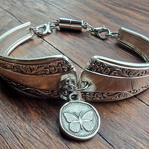 May include: A silver-toned bracelet crafted from a repurposed spoon handle featuring an ornate embossed pattern. The bracelet includes a circular charm with a butterfly design and a magnetic clasp connected by a small metal chain link.