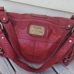 May include: A red handbag with a textured finish, featuring a top zip closure, brass-toned metal rings, and studs. A rectangular brass-toned metal plate on the front displays the text FOSSIL long live vintage since 1954 in embossed lettering.