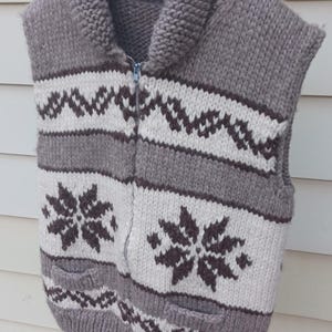 May include: A sleeveless knit vest featuring a full-length front zipper and a thick collar. The garment displays a pattern of dark brown snowflake motifs and horizontal zigzag stripes against a cream and light brown background.