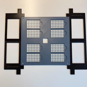 May include: A gray square grid plate with eight rectangular cutouts, flanked by two black rectangular frames labeled XTOOL M2 LARGE FRAME. The gray plate features a central square hole and a grid pattern, while the black frames have two rectangular openings each.