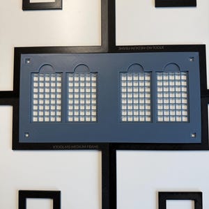 Może przedstawiać: Czarna ramka XTOOL-M2-MEDIUM-FRAME z geometrycznym, zazębiającym się wzorem obramowania otaczającym centralną niebieską prostokątną wkładkę. Niebieska wkładka posiada cztery łukowate wycięcia okienne z siatką i małe kwadratowe otwory.