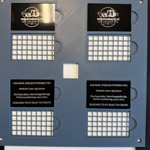 May include: A gray modular laser jig system base plate featuring four rectangular cutouts, each containing a black insert. The inserts display the text "mijnmakerij.nl" and "ONE BASE. ENDLESS POSSIBILITIES. Modular Laser Jig System."