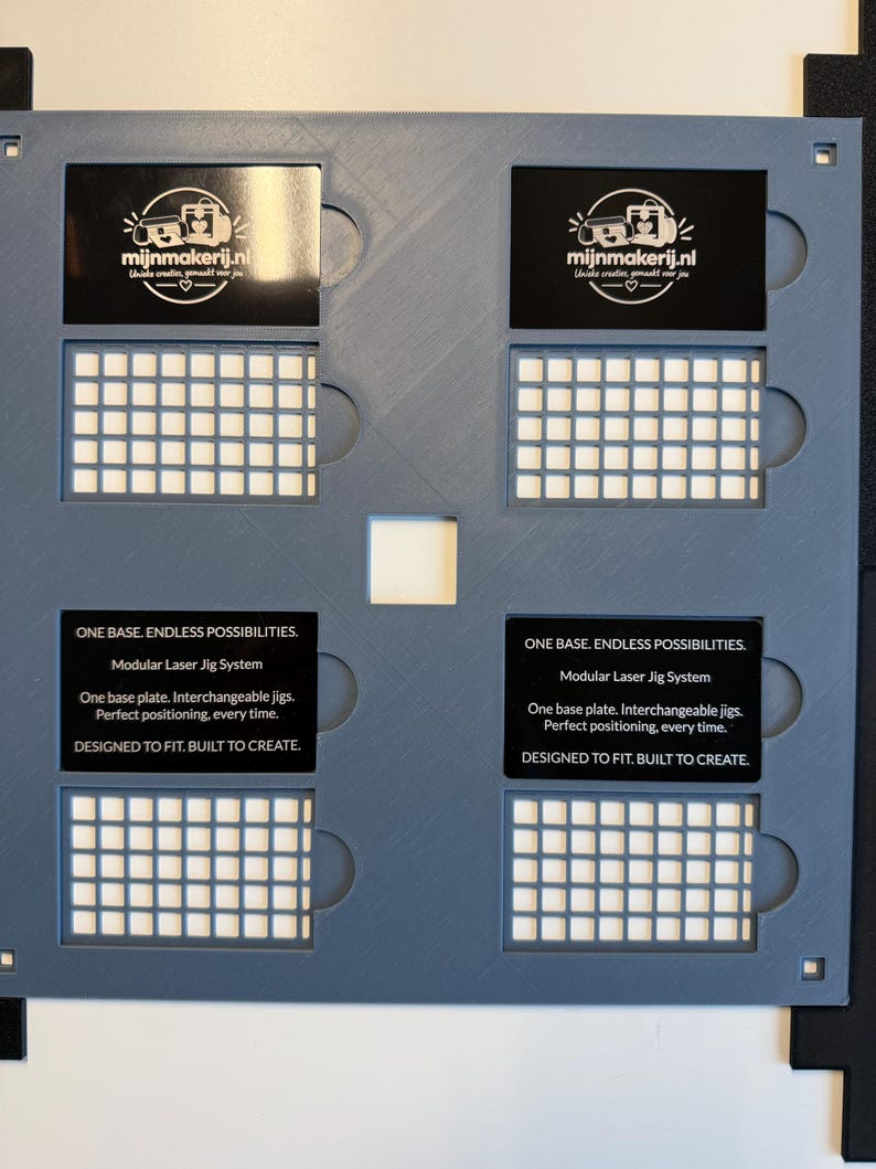 May include: A gray modular laser jig system base plate featuring four rectangular cutouts, each containing a black insert. The inserts display the text "mijnmakerij.nl" and "ONE BASE. ENDLESS POSSIBILITIES. Modular Laser Jig System."