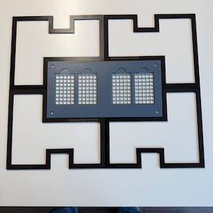 May include: A black xTool M2 medium frame consisting of four interlocking pieces arranged in a rectangular shape. Centered within the frame is a gray rectangular plate featuring four grid-patterned windows and small circular holes at the corners.