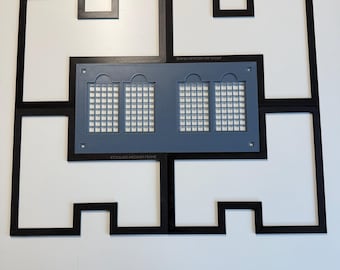 May include: A black xTool M2 medium frame consisting of four interlocking pieces arranged in a rectangular shape. Centered within the frame is a gray rectangular plate featuring four grid-patterned windows and small circular holes at the corners.