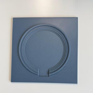 May include: A square, slate-blue plastic plaque featuring a raised, concentric circular design. The circular pattern is interrupted at the bottom by a small, rectangular cutout that extends from the inner ring to the edge of the square base.