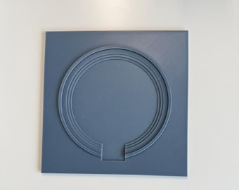 May include: A square, slate-blue plastic plaque featuring a raised, concentric circular design. The circular pattern is interrupted at the bottom by a small, rectangular cutout that extends from the inner ring to the edge of the square base.