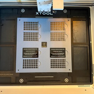 May include: A black xTool laser machine base plate featuring a central light gray modular jig system with grid patterns. The plate includes two black labels with white text reading: ONE BASE. ENDLESS POSSIBILITIES. Modular Laser Jig System.