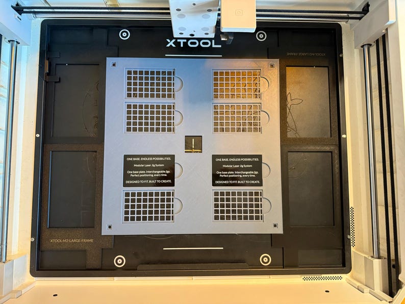 May include: A black xTool laser machine base plate featuring a central light gray modular jig system with grid patterns. The plate includes two black labels with white text reading: ONE BASE. ENDLESS POSSIBILITIES. Modular Laser Jig System.