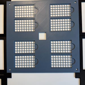 May include: A square, slate-blue plastic grid template featuring six rectangular cutouts with internal mesh patterns and small circular side tabs. A small square hole is centered in the middle, with four black corner frames visible.