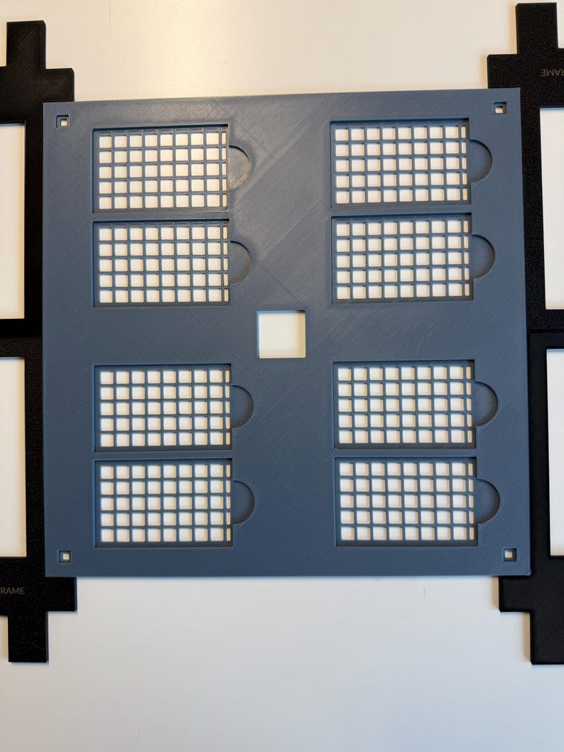 May include: A square, slate-blue plastic grid template featuring six rectangular cutouts with internal mesh patterns and small circular side tabs. A small square hole is centered in the middle, with four black corner frames visible.