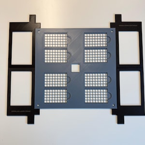 May include: A gray square grid plate with eight rectangular cutouts, flanked by two black rectangular frames labeled XTOOL M2 LARGE FRAME. The gray plate features a central square hole and a grid pattern, while the black frames have two rectangular openings each.