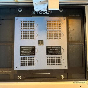 May include: A light gray modular laser jig system plate with four grid sections and black labels reading ONE BASE, ENDLESS POSSIBILITIES. Modular Laser Jig System. One base plate. Interchangeable jigs. Perfect positioning, every time. DESIGNED TO FIT. BUILT TO CREATE.