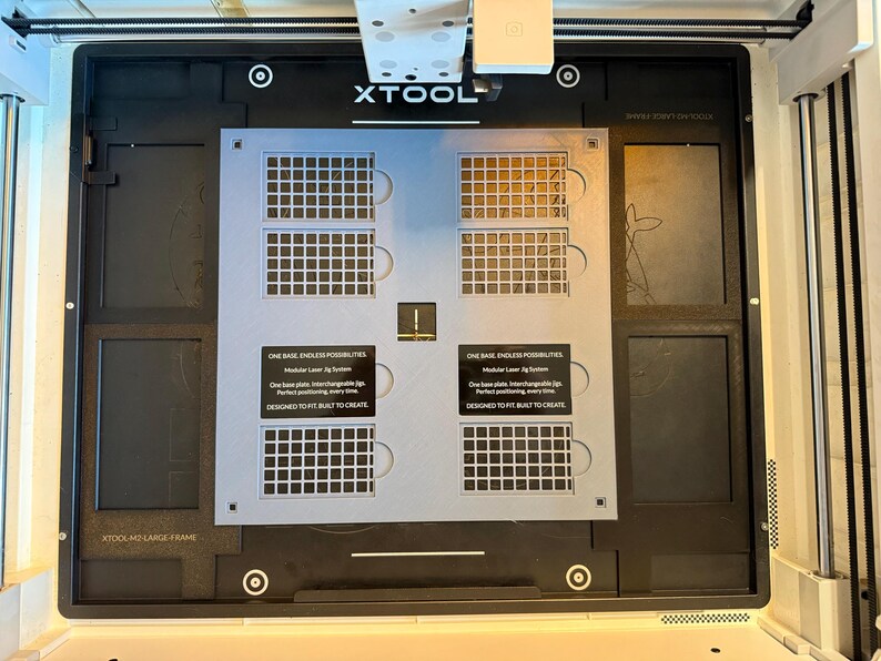 May include: A light gray modular laser jig system plate with four grid sections and black labels reading ONE BASE, ENDLESS POSSIBILITIES. Modular Laser Jig System. One base plate. Interchangeable jigs. Perfect positioning, every time. DESIGNED TO FIT. BUILT TO CREATE.