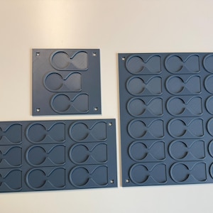 May include: Three dark gray rectangular storage organizers with multiple recessed, figure-eight shaped slots. The organizers feature small square mounting holes in each corner, designed for holding small items in a grid-like arrangement.