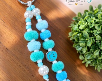 Peut inclure: Un porte-clés composé de deux brins de perles attachés à un anneau en métal argenté. Les brins incluent des perles en plastique blanc et bleu clair, des perles nacrées, des pompons bleu et turquoise, et un jeton bleu avec un visage souriant.