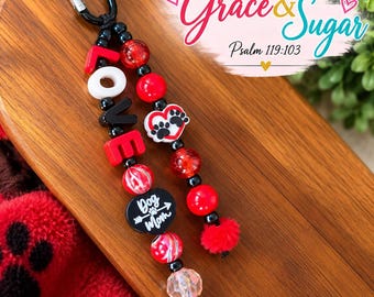 Peut inclure: Un porte-clés perlé avec un mousqueton noir, des perles rouges et noires, et des lettres formant le mot LOVE. Il comprend une breloque circulaire noire avec le texte Dog Mom et une flèche, un cœur blanc avec des empreintes de pattes et des pompons rouges.