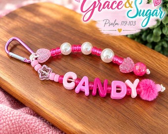 Peut inclure: Un porte-clés en perles avec un mousqueton violet, des perles roses et blanches, une perle en forme d'étoile, deux cœurs pailletés et des lettres formant le mot CANDY, terminé par un pompon rose. Le texte Grace & Sugar Psalm 119:103 est visible.