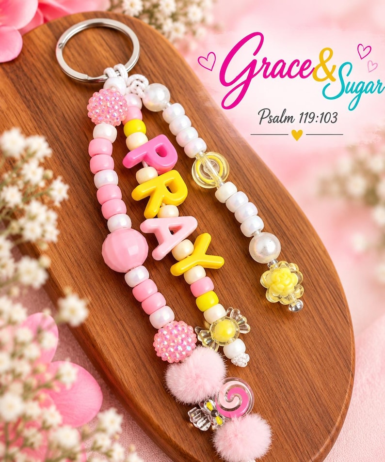 Peut inclure: Un porte-cl&eacute;s compos&eacute; de deux brins de perles color&eacute;es, incluant des sph&egrave;res roses, jaunes et blanches, avec le mot PRAY &eacute;pel&eacute; en lettres majuscules jaunes et roses. Le design comprend des petits pompons et des breloques en forme de bonbons, avec le texte Grace & Sugar Psalm 119:103 visible dans le coin sup&eacute;rieur droit.