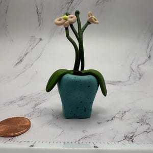 May include: A miniature, handcrafted polymer clay orchid plant in a turquoise pot. The plant features green leaves and stems with delicate, light peach-coloured flowers. A penny is next to the plant for scale. The pot has a speckled texture.