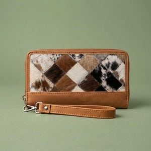 May include: A rectangular wallet featuring a patchwork design of brown, black, and white cowhide squares on the front panel. The wallet is constructed with tan leather trim and includes a matching detachable tan leather wrist strap.