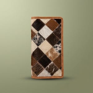 May include: A rectangular wallet featuring a patchwork design of cowhide squares in shades of brown, black, white, and tan. The wallet is framed by a smooth tan leather border, creating a structured appearance with a visible hair-on texture.