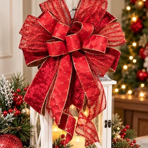 May include: A large, multi-layered decorative bow in deep red, featuring a mix of sheer, glittery striped ribbon and solid red ribbon with a subtle diamond-patterned texture. The bow has long, flowing tails and gold-trimmed edges.