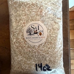 May include: A clear plastic resealable bag filled with crushed off-white eggshells. A circular white label features a tree, a chicken, and a beehive illustration with the text S&I ROOT, ROOST & HIVE LLC. The bag is marked with 14oz in black ink.
