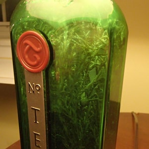 May include: A green glass bottle with a red wax seal and a metal label that reads "No. I E". The bottle contains green plant matter. The bottle is a decorative item.