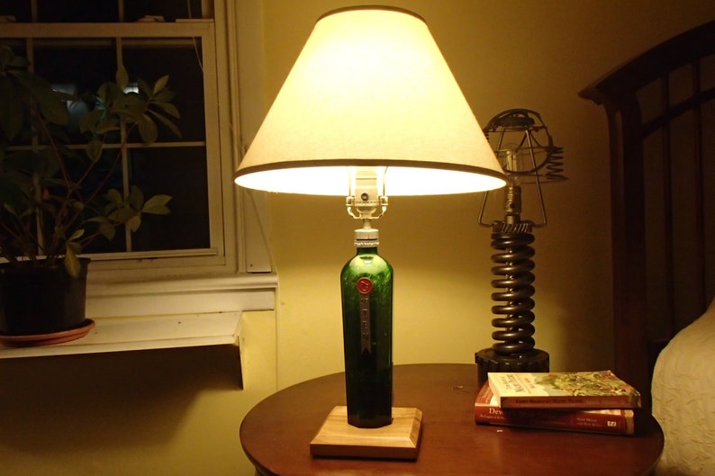 May include: A unique table lamp crafted from a green bottle, featuring a beige lampshade. The lamp is mounted on a wooden base and sits on a round, dark brown table. A second, industrial-style lamp is also visible.