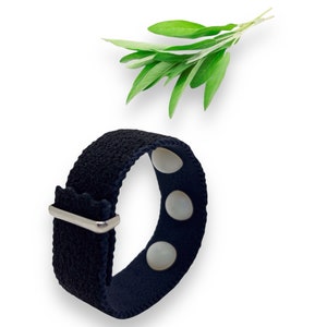 May include: A navy blue wristband with three white buttons and a silver-colored metal clasp. Fresh green sage leaves are positioned above the wristband against a white background. The wristband is made of a textured fabric.