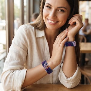 May include: Two woven bracelets in shades of purple, blue, and tan are worn on the wrists. The bracelets have a textured appearance and a mix of colours. The person is wearing a white button-down shirt.