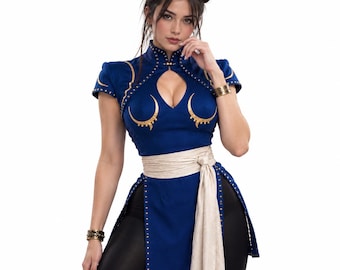 May include: A blue qipao-style top featuring a high collar, short sleeves, and a keyhole neckline. The garment is adorned with gold crescent-shaped accents and gold stud detailing along the edges. A wide, light-colored sash is tied at the waist.