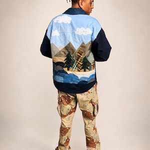 May include: A long-sleeve button-up shirt featuring a patchwork landscape design on the back with light blue sky, white clouds, brown mountains, green pine trees, and blue water. The shirt has dark navy sleeves and collar. Paired with camouflage cargo pants.