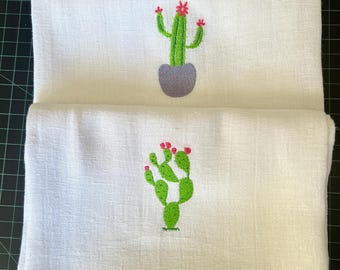 Puede incluir: Dos toallas de mano blancas con diseños de cactus bordados. Una toalla presenta un cactus verde en una maceta gris con flores rojas. La otra toalla tiene un cactus verde con flores rojas. Las toallas están dobladas y apiladas.