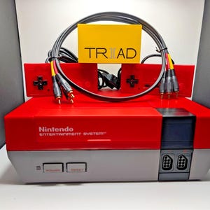 以下が含まれることがあります： 赤とグレーのNintendo Entertainment System本体、2つの赤いコントローラー、黒いAVケーブル、TRIADと書かれた黄色い箱。本体にはフロントローディングスロット、電源ボタン、リセットボタン、2つのコントローラーポートが備わっています。