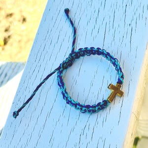 May include: A braided cord bracelet featuring a mix of dark blue and light blue threads. The bracelet includes a small, metallic gold-toned cross charm integrated into the woven design, with two loose cord ends extending from the adjustable knot.