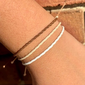 Puede incluir: Un conjunto de tres pulseras finas de cordón trenzado en tonos marrón, beige y blanco. Cada pulsera presenta un diseño trenzado sencillo hecho de hilo fino, que se lleva junto como una pila en capas en la muñeca con extremos ajustables.