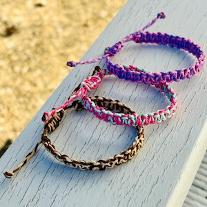 May include: Three adjustable braided cord bracelets. The top bracelet features purple and blue tones. The middle bracelet is woven with bright pink and light blue threads. The bottom bracelet is crafted from brown and cream colored cord.