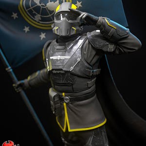 Pode incluir: Uma figura de soldado com armadura tática cinza-escura e preta com detalhes amarelos, usando um capacete com viseira, saudando enquanto segura uma grande bandeira azul-escura com um emblema circular e estrelas brancas ao fundo.