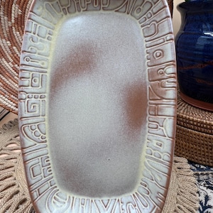 May include: A rectangular ceramic serving tray with rounded corners, featuring a smooth, light beige centre with subtle brown shading and a wide, textured border decorated with an intricate, embossed geometric pattern in tan and cream tones.