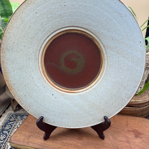 May include: A round ceramic plate featuring a wide, speckled off-white rim and a deep, solid terracotta-colored center. The center of the plate displays a subtle, darker spiral pattern. The plate is supported by a dark brown plastic display stand.