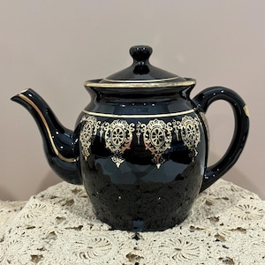 May include: A glossy black ceramic teapot featuring a gold-colored decorative pattern around the upper body. The teapot includes a matching lid with a small knob handle, a curved spout with gold trim, and a sturdy handle on the side.